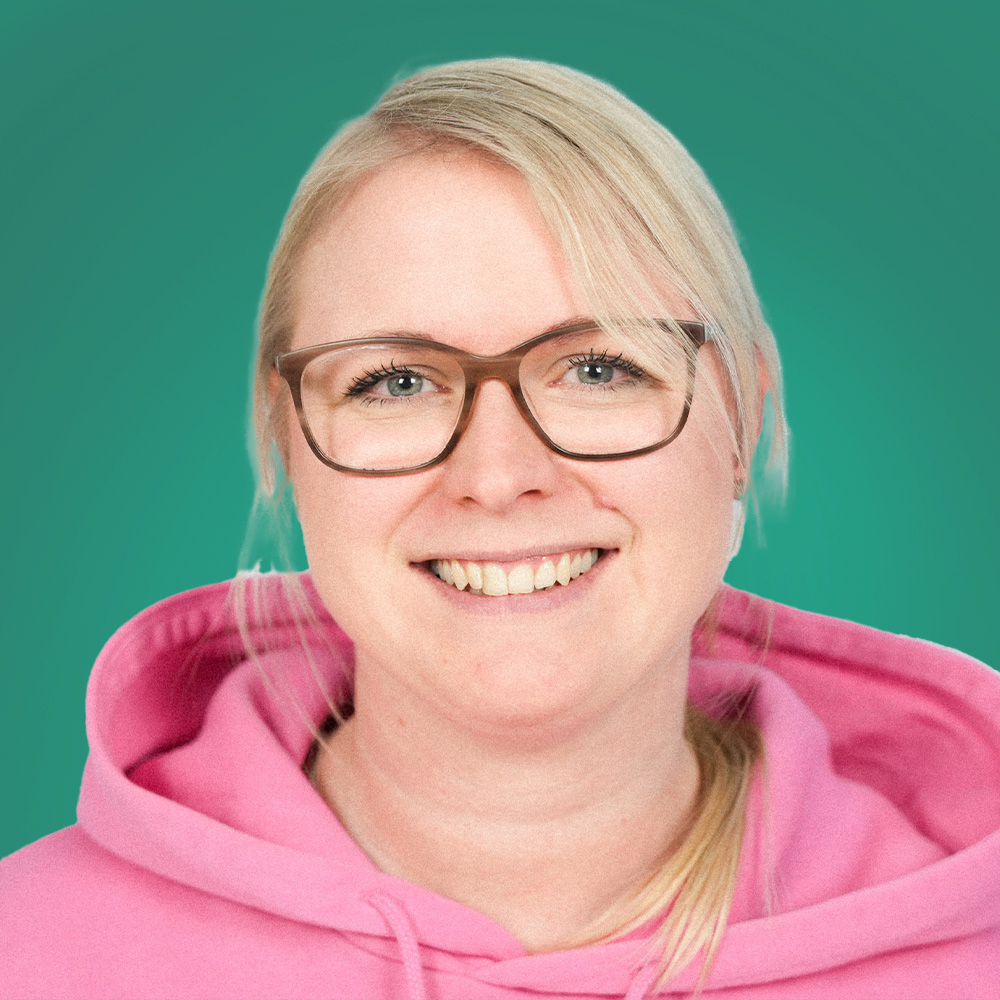 Porträt einer Person mit heller, schulterlanger Frisur und Brille vor türkisem Hintergrund, rosa Pullover